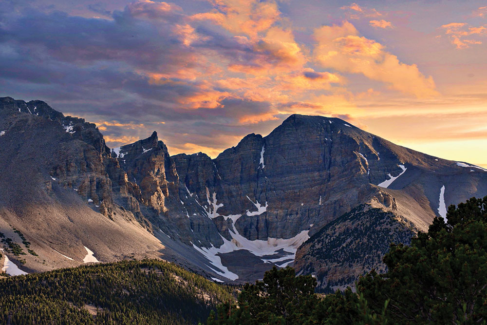Mountain with snow patches, illuminated by sunset, under colorful sky, surrounded by dense green forest.